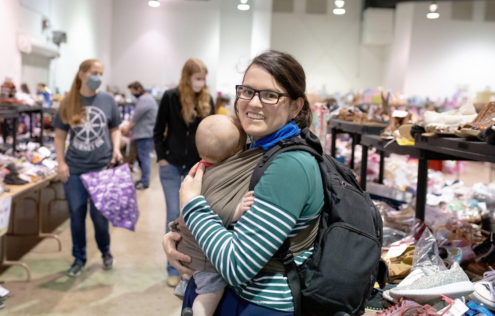 Three generations shop together at their local JBF sale. Mom carries baby in a baby carrier while grandmom stands behind them.