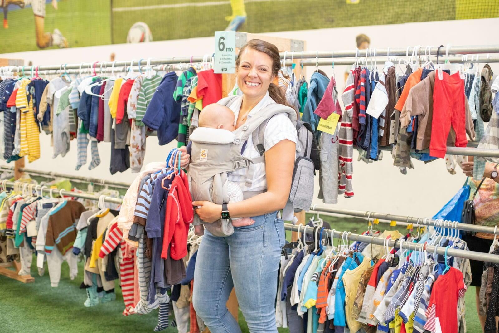 Three generations shop together at their local JBF sale. Mom carries baby in a baby carrier while grandmom stands behind them.