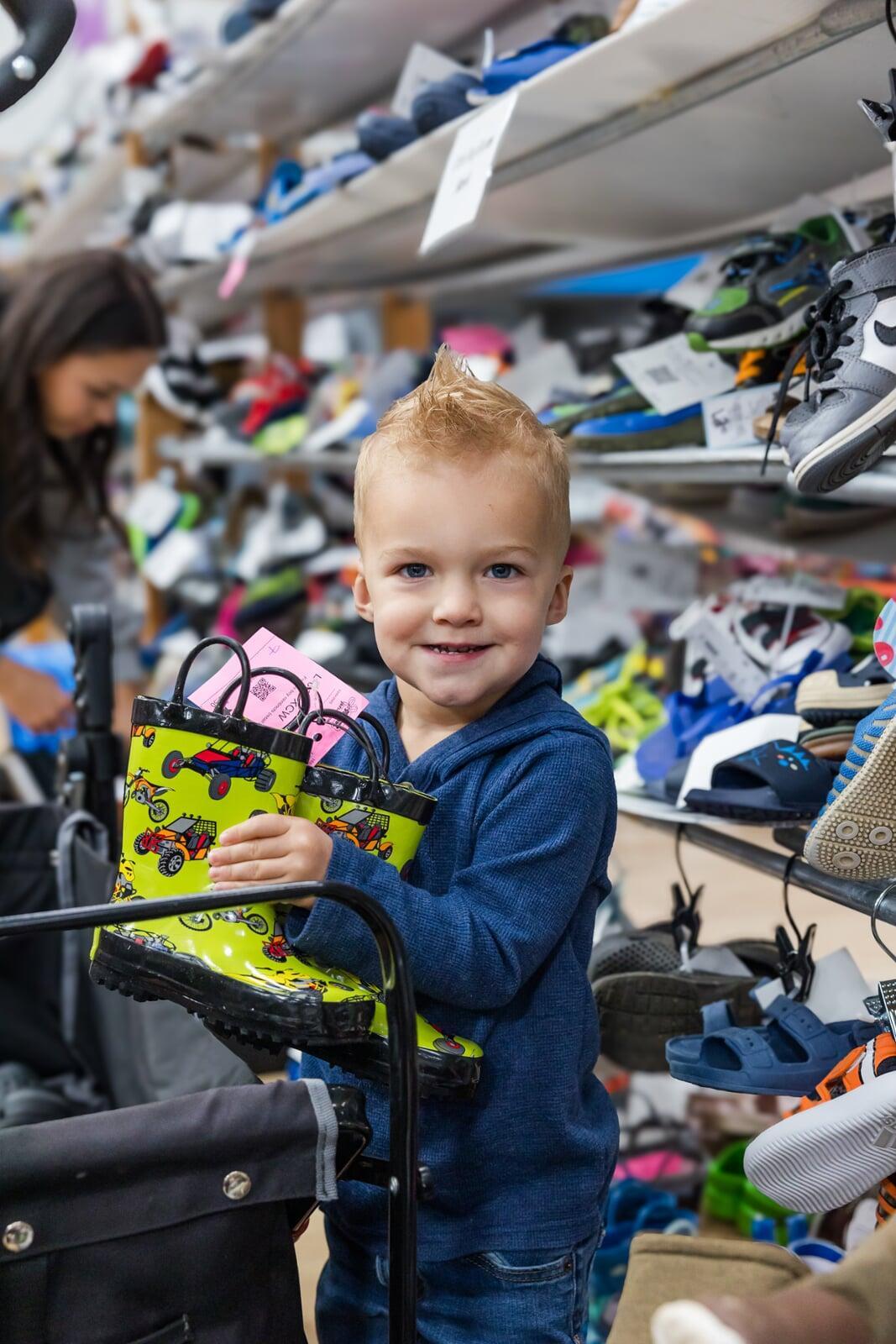 A mom with a large JBF shopping bag on her shoulder stands beside her husband who wears their toddler at a JBF sale.